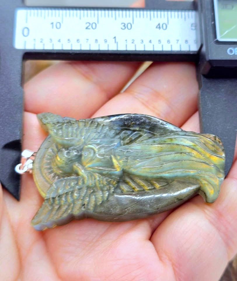 A hand holds a carved pendant or charm shaped like a stylized bird or fish, shown under a digital caliper for size reference. The item appears olive green to gray with yellow-gold highlights and a small silver ring at the top.