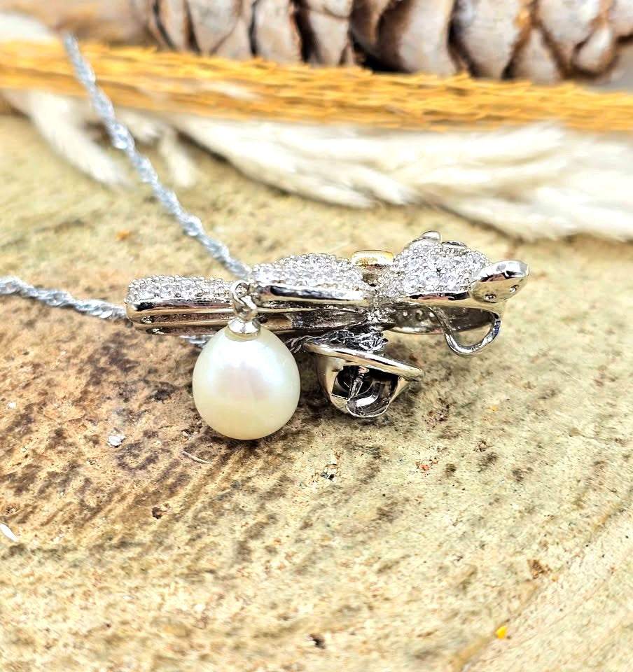 A teddy bear pendant covered in sparkling clear stones, wearing a gold bow tie, with a silver chain and a white pearl hanging from one hand in side view-My Aura Gemstones
