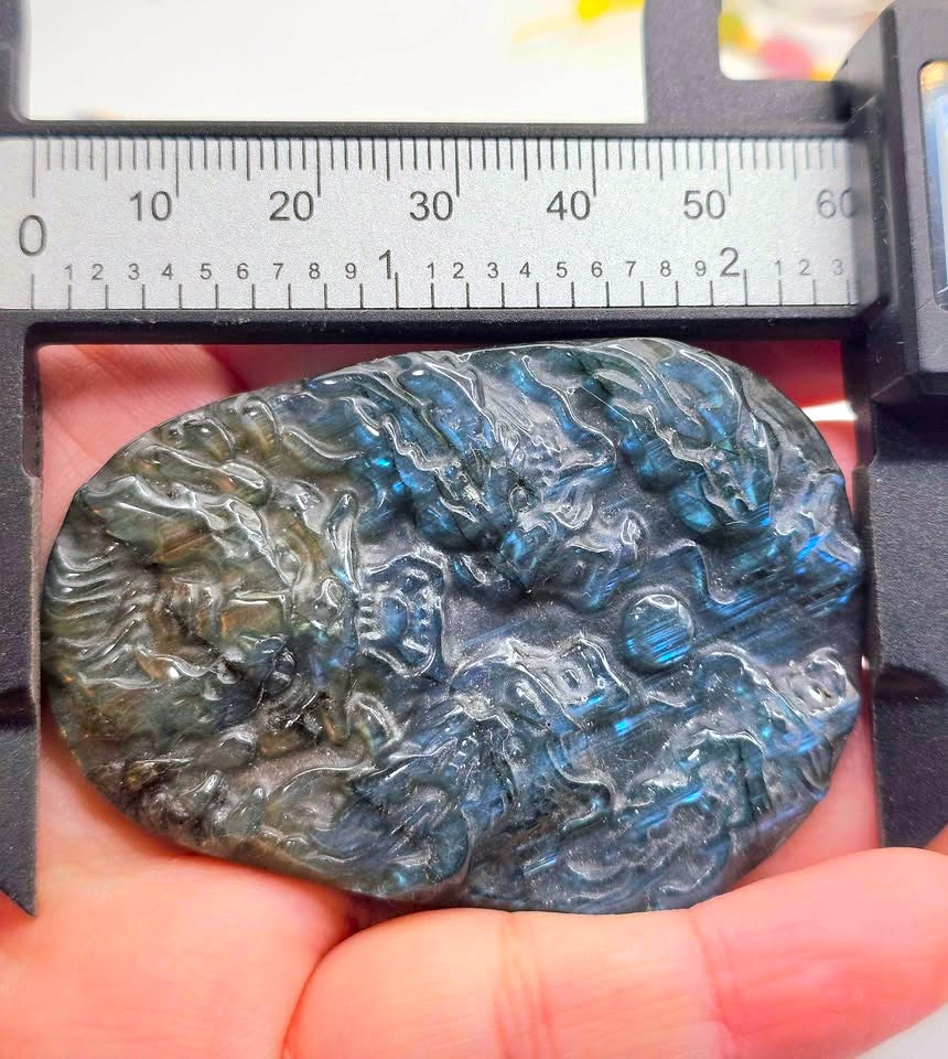 A hand holds a polished irregular stone pendant with a blue-gray labradorite finish and glittering flashes, positioned under a metal caliper with a ruler scale visible at the top for size reference.