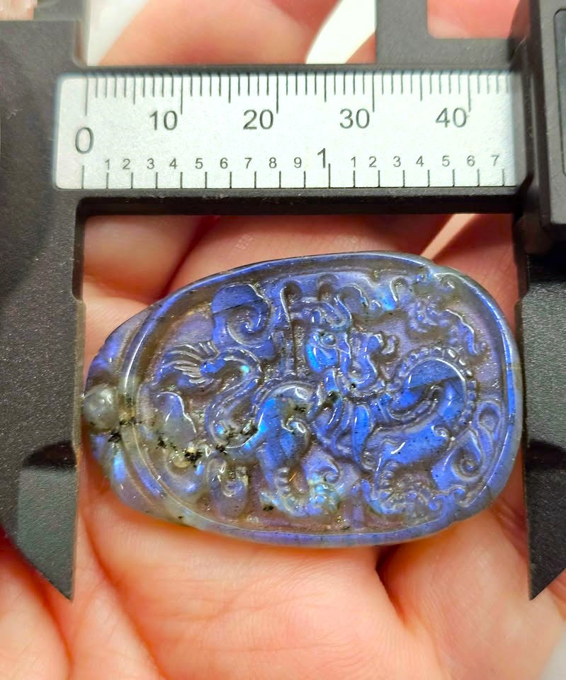 A close-up of a carved oval pendant or charm with an iridescent blue-purple labradorite-like surface, held in a hand under a caliper for scale. The pendant has raised decorative relief patterns and is photographed against skin with a measuring tool visible at the top.