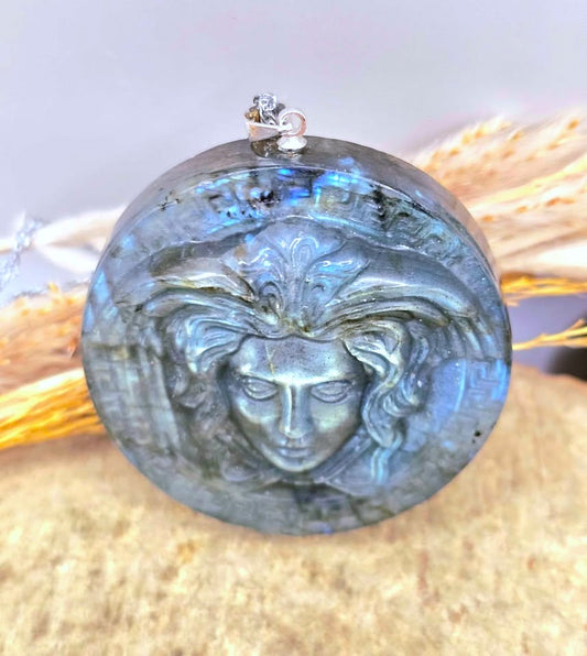 A round labradorite gemstone pendant featuring a raised Medusa-style face motif, with a silver bail and hanging loop at the top, displayed against a neutral background with dried wheat stalks.