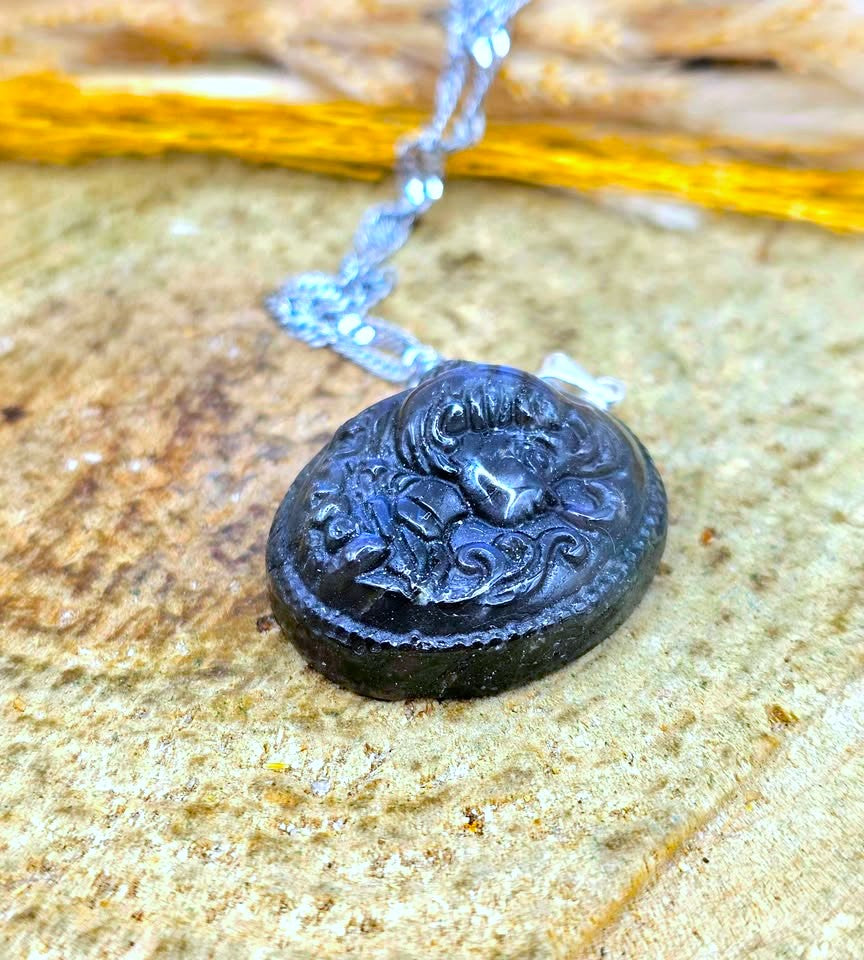 A dark carved pendant necklace with a silver-toned chain, resting on a textured stone surface.