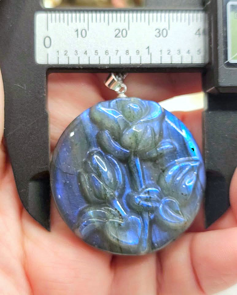 A round labradorite pendant with a carved floral relief, held in a hand under a caliper to show its size. The pendant exhibits a blue-gray color with a strong greenish iridescent flash.