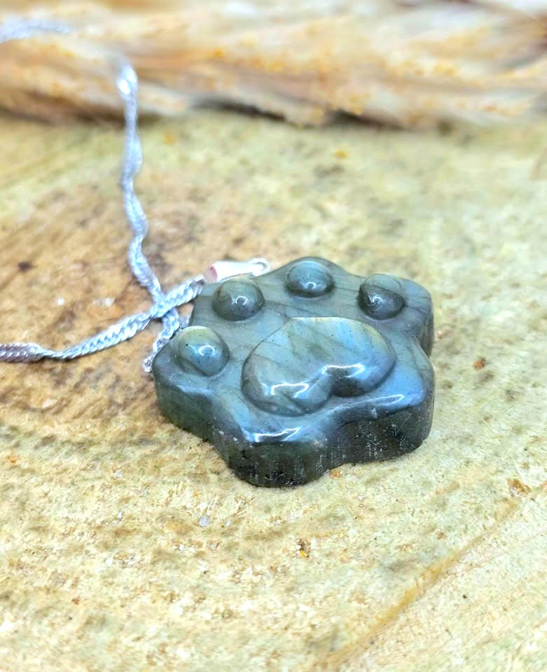 A polished dark gray paw print pendant necklace on a silver chain, resting on a textured sandy surface.