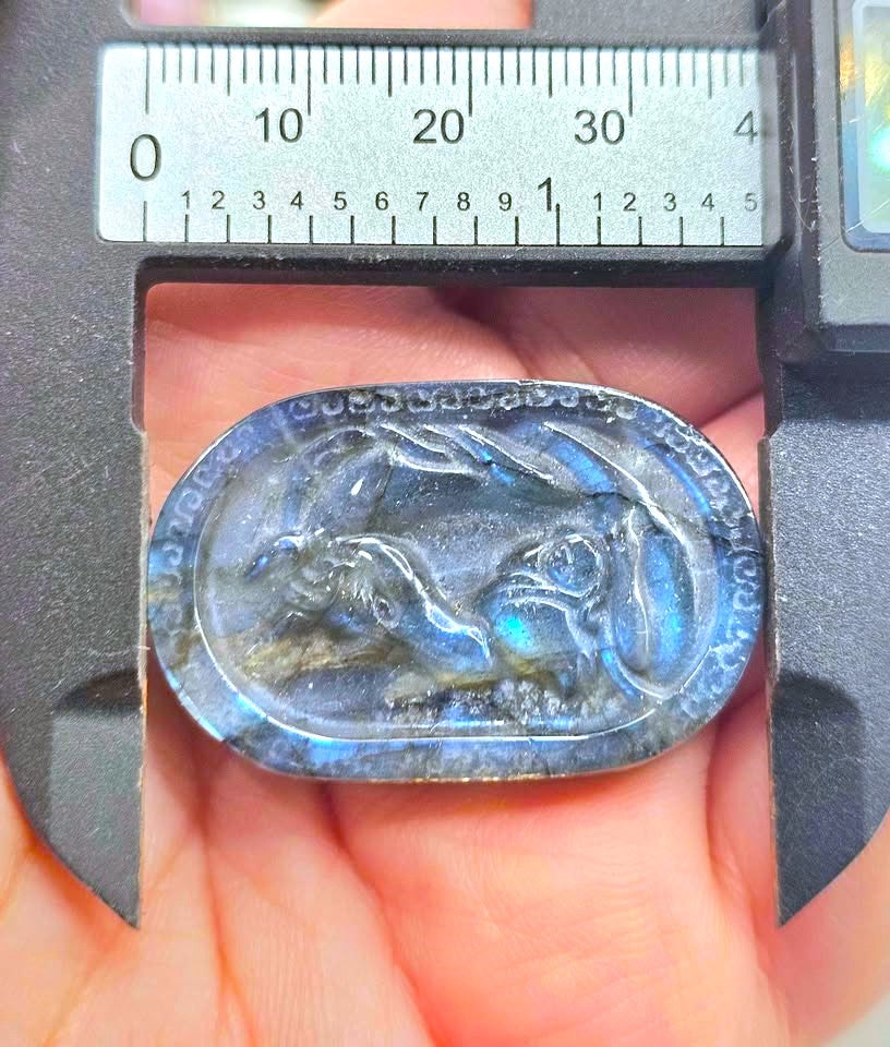A close-up of a hand holding an oval labradorite pendant under a digital caliper to show its size. The pendant features a carved rabbit design with a blue iridescent labradorite finish.