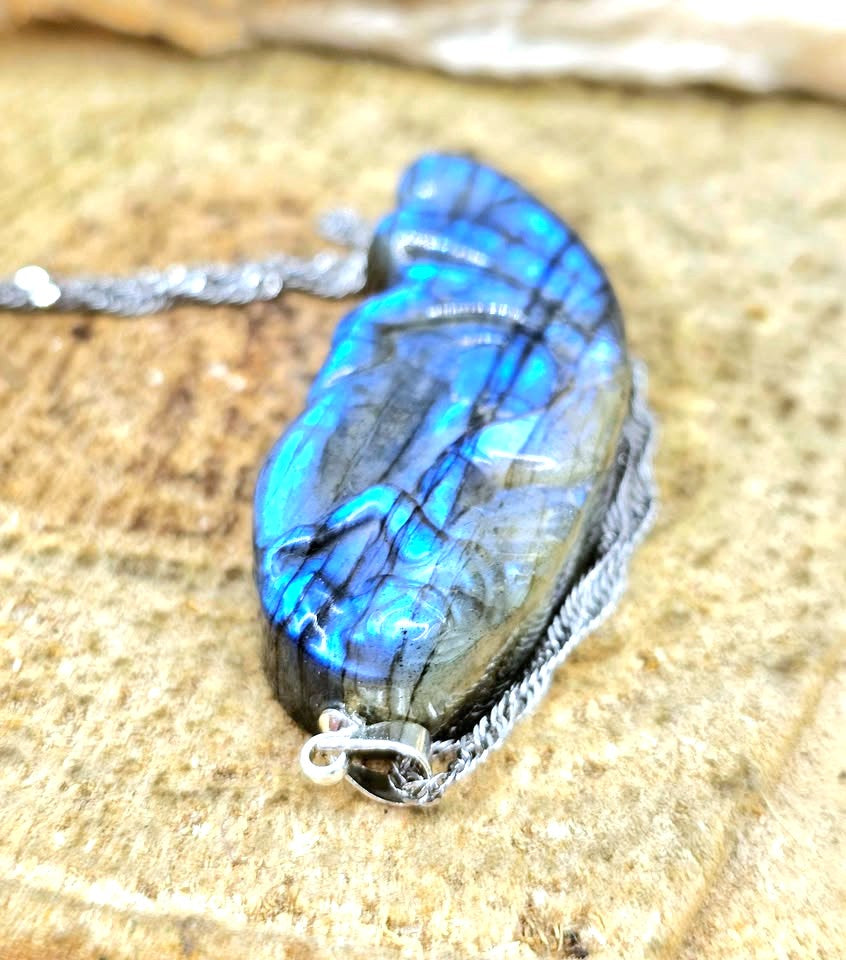A silver chain necklace with a polished labradorite pendant, resting on a textured stone surface. The pendant displays vibrant blue and green iridescent flashes with dark gray veining.