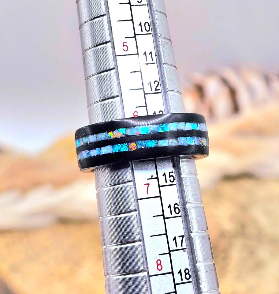 A black ring with a vibrant blue and orange inlay, shown on a silver metal ring sizer against a blurred desert background.