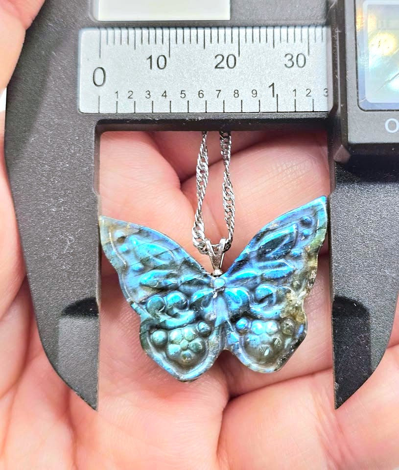 A close-up of a butterfly-shaped pendant with iridescent blue and green labradorite wings, held in a hand under a digital caliper to show its size. The pendant hangs from a silver chain.