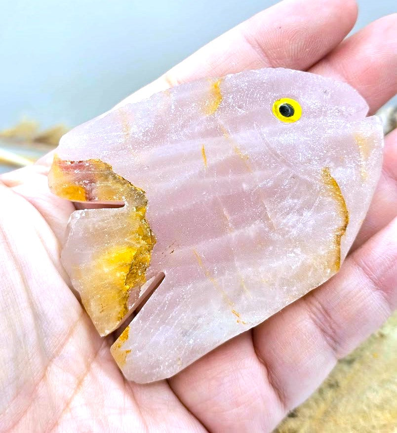 A hand holding a carved pale pink stone fish figurine with yellow and orange mineral accents and a small yellow eye.