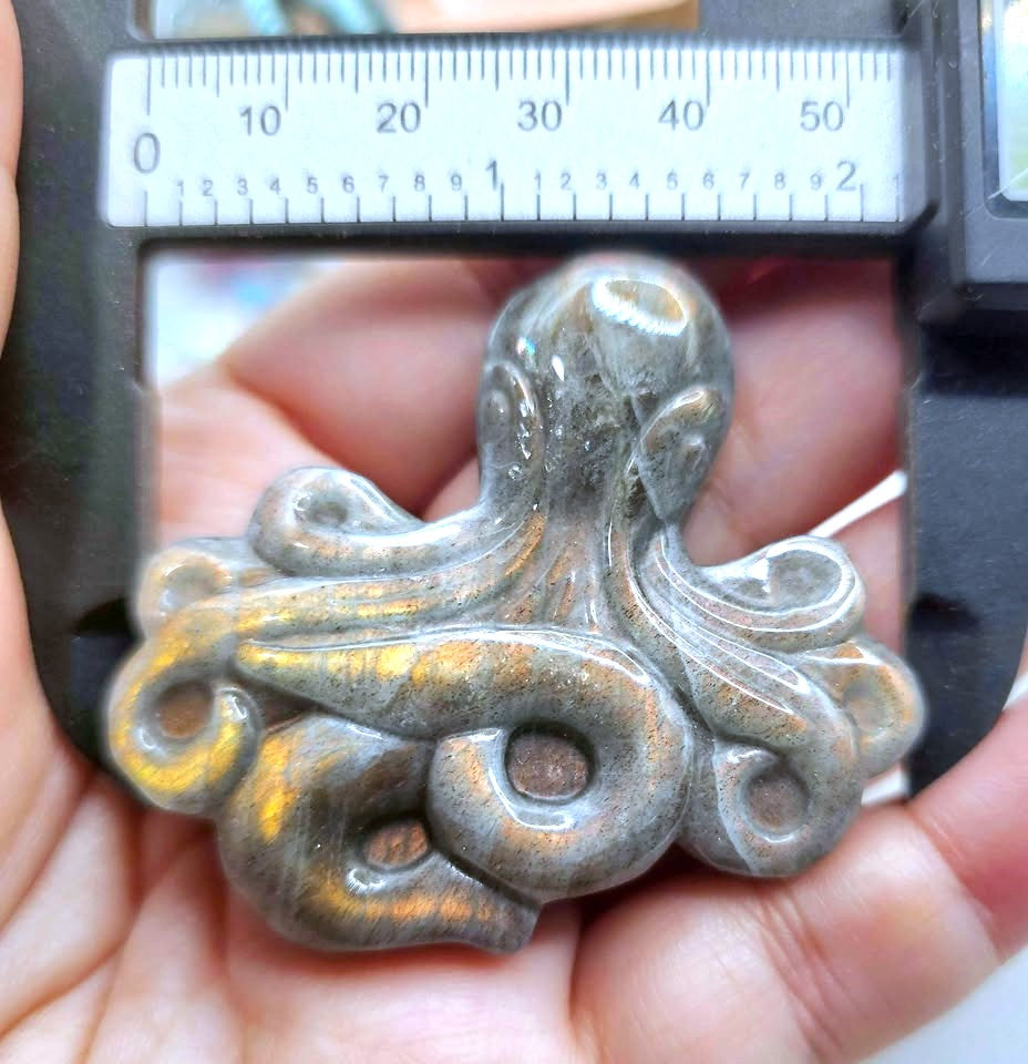 A carved octopus-shaped pendant or charm in a metallic silver-gray finish, held in a person's hand under a caliper to show scale.