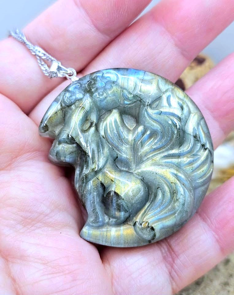 A close-up of a hand holding a carved oval pendant featuring a stylized animal head, likely a wolf or fox, with a silver-toned chain. The pendant has a gray and gold marbled pattern with natural stone veining.