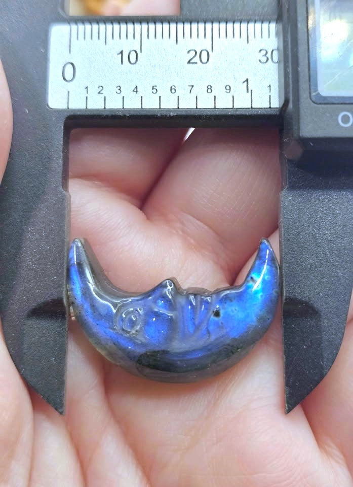 A close-up of a crescent moon-shaped cabochon with a vibrant blue iridescent labradorescent finish, held in a person's hand under a black caliper to show its size.