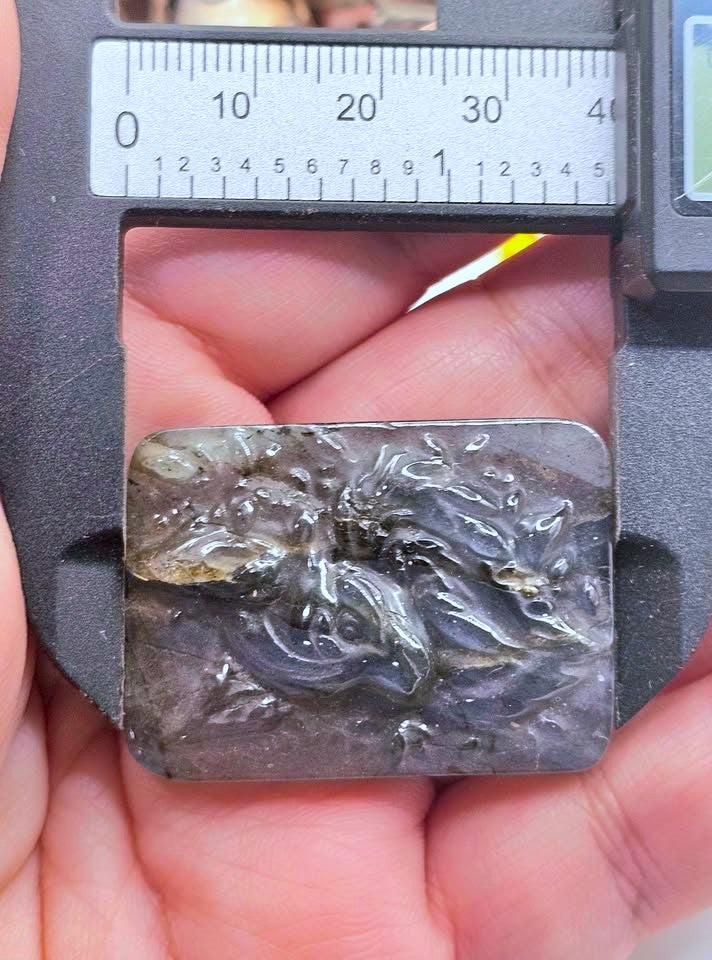 A hand holding a carved rectangular stone slab under a digital caliper to show its dimensions. The stone has a dark gray to black base with prominent swirling white and light gray marbled patterns, resembling a wood or stone carving.