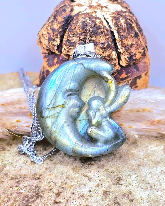 A silver pendant necklace featuring a carved labradorite crescent moon and baby figurine, displayed on a textured stone surface with a dried seed pod in the background.