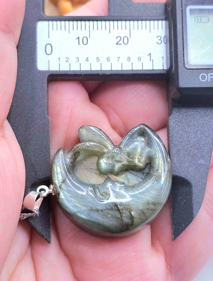 A polished labradorite pendant shaped like a crescent moon with a rabbit motif, held in a hand under a digital caliper to show its size.