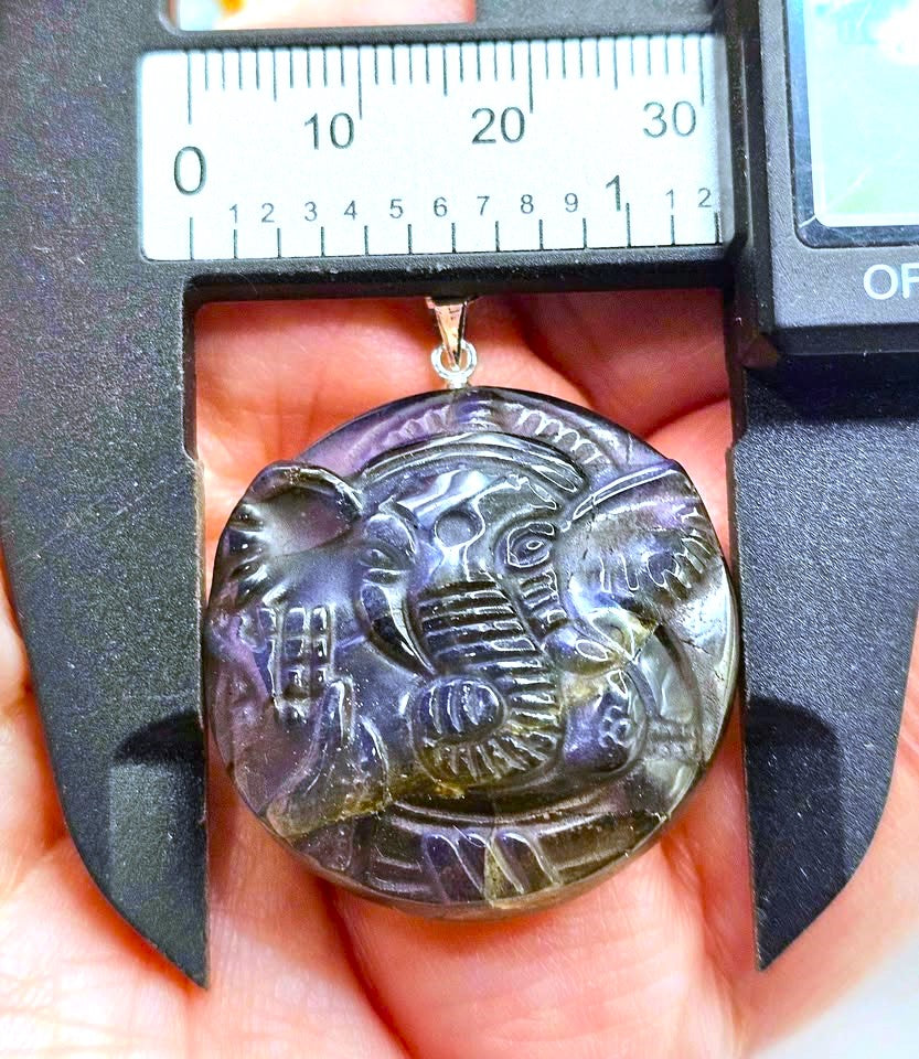 A round carved pendant with an elephant design, held in a hand under a digital caliper to show its size. The pendant has a dark, smoky translucent appearance with purple and green iridescent tones.