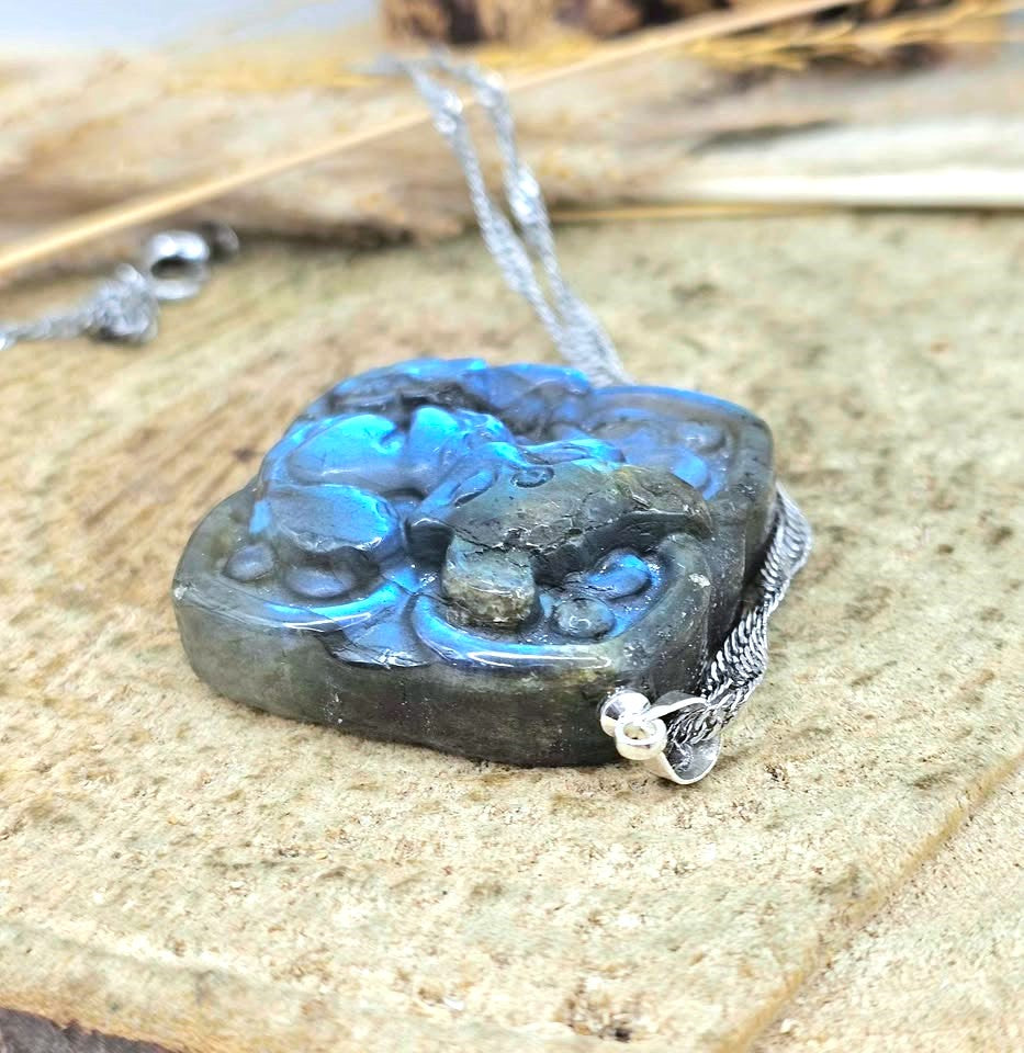 A carved stone pendant necklace with a silver chain, featuring a dark gray stone with vibrant blue iridescent inclusions, resting on a textured stone surface.