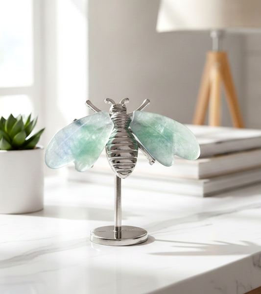 A silver metal bee figurine with natural fluorite wings, mounted on a round silver base and displayed on a textured sandy surface display in indoor room ondesk - My Aura Gemstones
