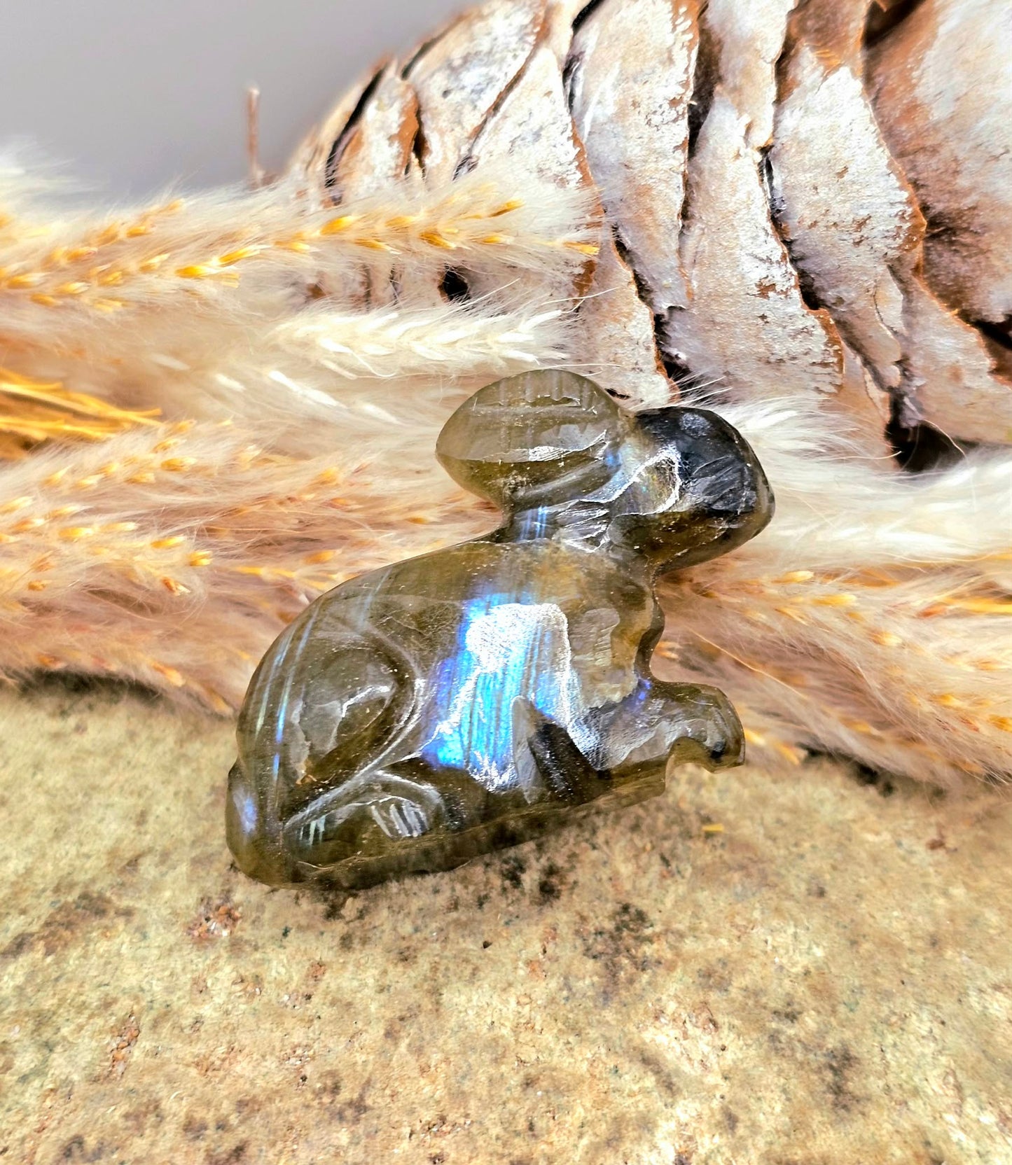 One  hand carved rabbit figurines displayed on a natural wood slice, with a pinecone and dried grass in the background.