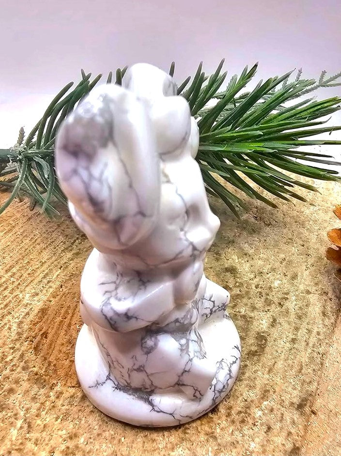 A white howlite rabbit figurine of a seated figure, styled on a sandy surface with green pine needles in the background.