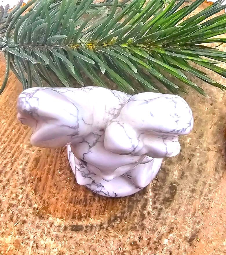 A white howlite rabbit figurine with gray veining, placed on a textured tan surface with green pine needles in the background.