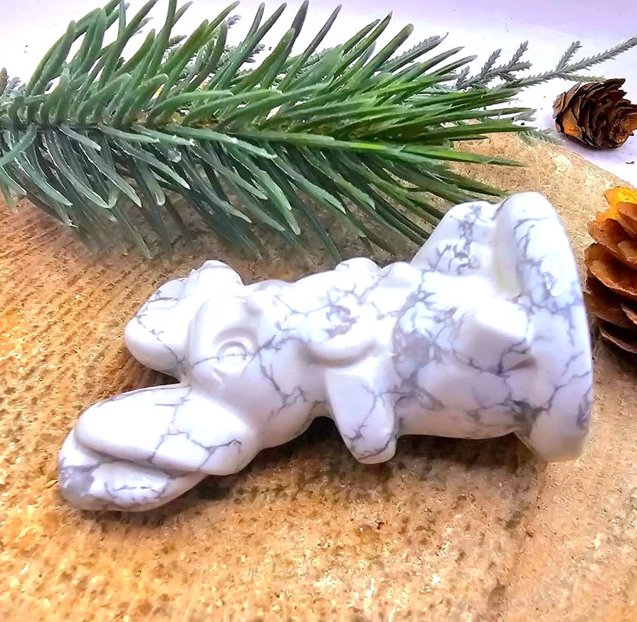 A white howlite carved stone rabbit figurine with gray veining, displayed on a textured tan surface with pine needles and pinecones in the background-My Aura Gemstones