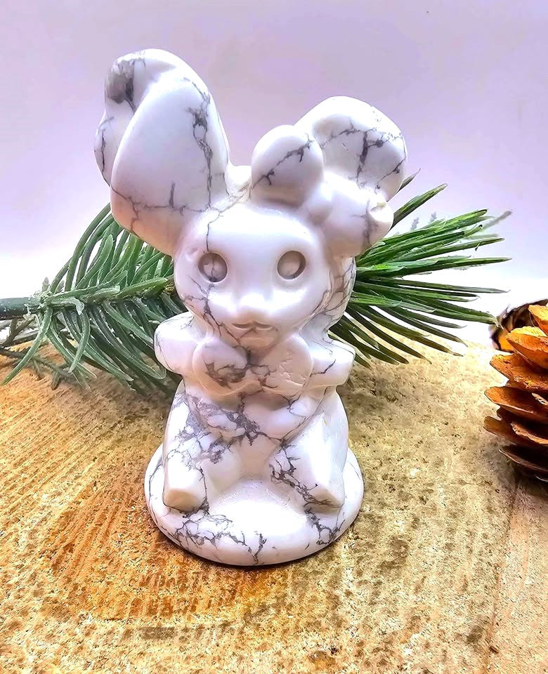A white howlite rabbit figurine with gray veining, sitting on a wooden surface with green pine branches behind it.
