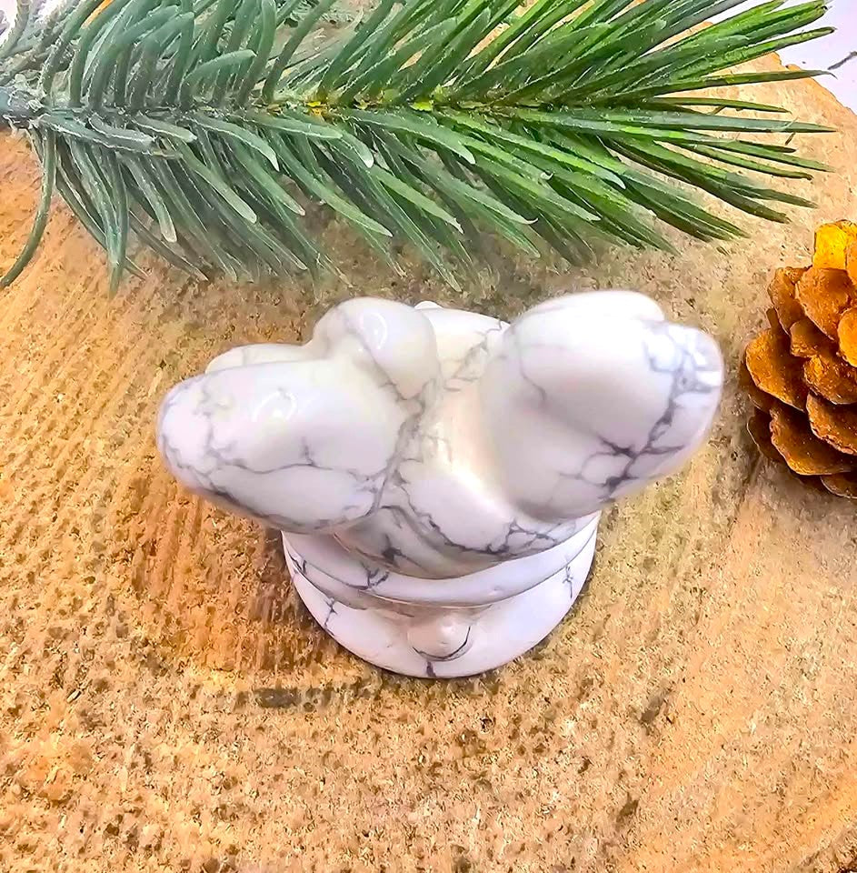 A white howlite figurine with gray veining, shaped like a stylized animal head, displayed on a textured tan surface with a green pine branch and a pinecone -My Aura Gemstones