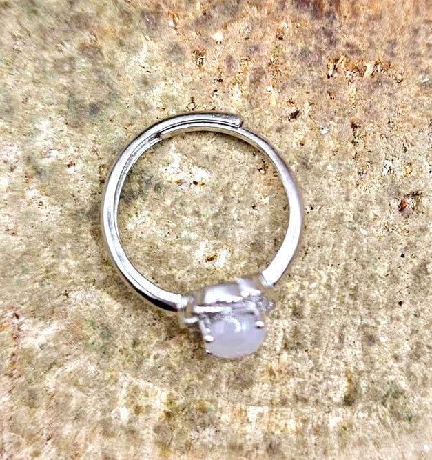 A silver-toned ring with a round, milky white gemstone, displayed on a textured beige stone surface.