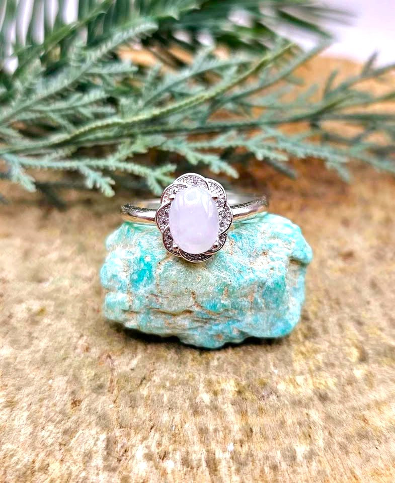 A silver ring with an oval pale lavender stone, displayed on a turquoise-colored rock with green pine branches in the background.