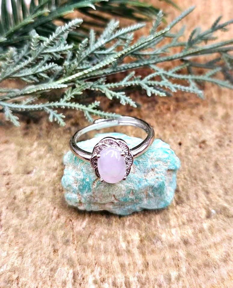 A silver-toned adjustable ring with an oval pale pink stone and small clear accent stones, displayed on a turquoise-colored rock with green foliage in the background.