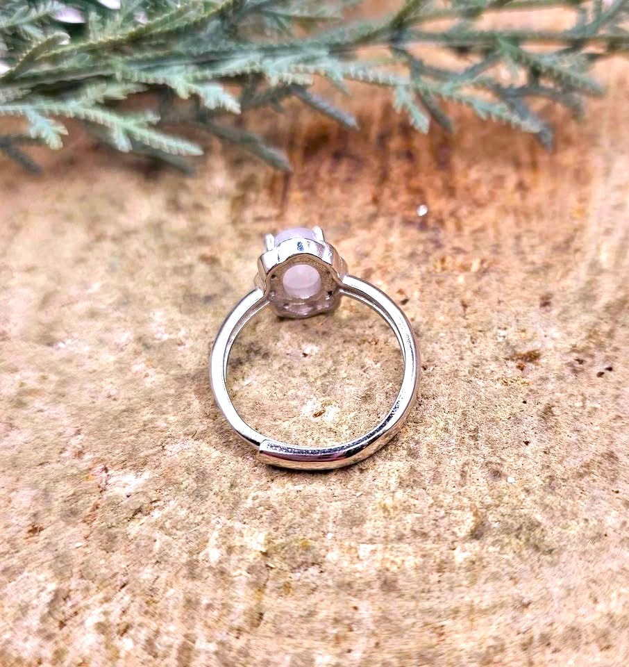 A silver-toned ring with a bezel-set pale pink gemstone, displayed on a textured tan stone surface with green foliage in the background.