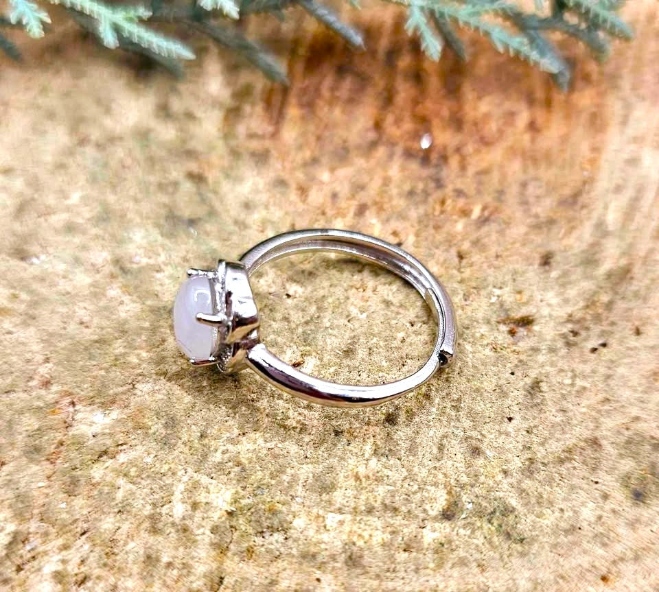 A silver-toned ring with a round, milky white cabochon gemstone, resting on a textured sandy surface with green foliage in the background.