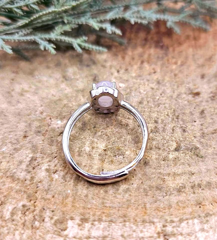 A silver-toned ring with a round, bezel-set pale pink stone, photographed on a textured tan stone surface with green foliage in the background.