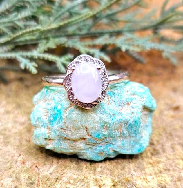 A silver ring with an oval pale lavender cabochon gemstone and a scalloped halo of small clear stones, displayed on a turquoise stone with green foliage in the background.