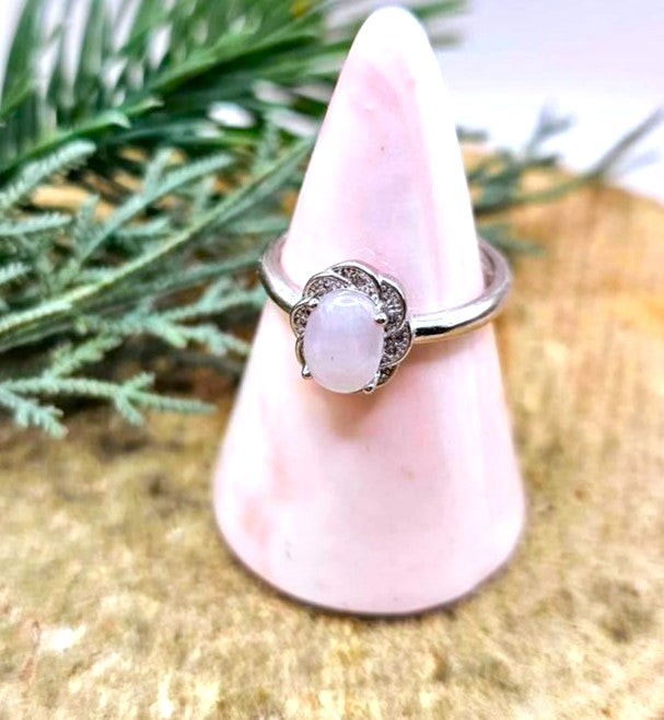 A silver ring with an oval pale pink stone and a halo of small clear stones, displayed on a pink cone stand with green foliage and a textured surface in the background.