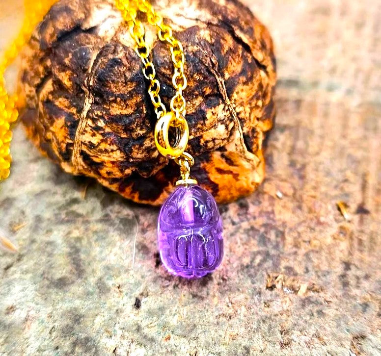 A close-up of a pendant necklace featuring a carved purple amethyst rabbit on a gold chain, resting on a textured brown wood chip in  front view-My Aura Gemstones