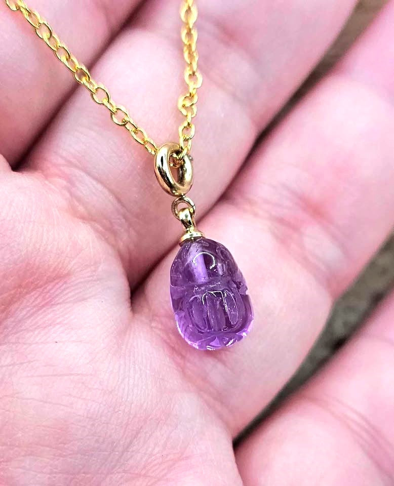 A close-up of a pendant necklace featuring a carved purple amethyst rabbit on a gold chain, resting on a textured brown wood chip-My Aura Gemstones