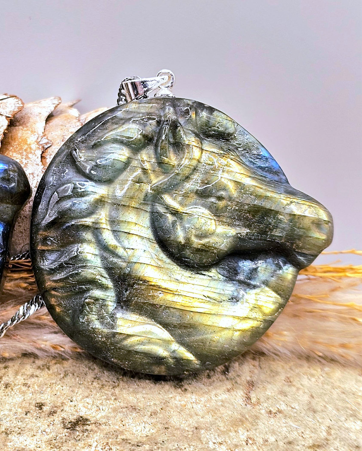 A round labradorite stone pendant carved into a horse head, resting on a textured tan surface with a silver bail at the top.