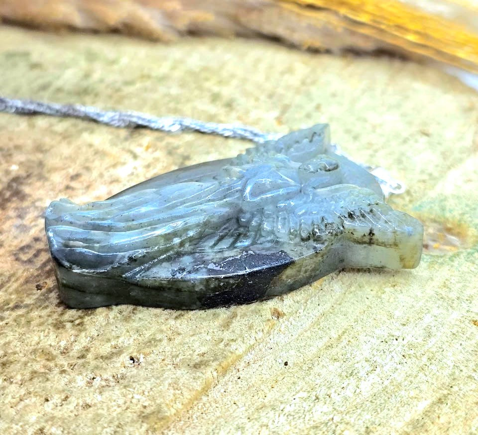 A carved stone pendant or charm with a dark gray to greenish-gray color, featuring a textured, organic shape, shown resting on a sandy surface with a silver chain.