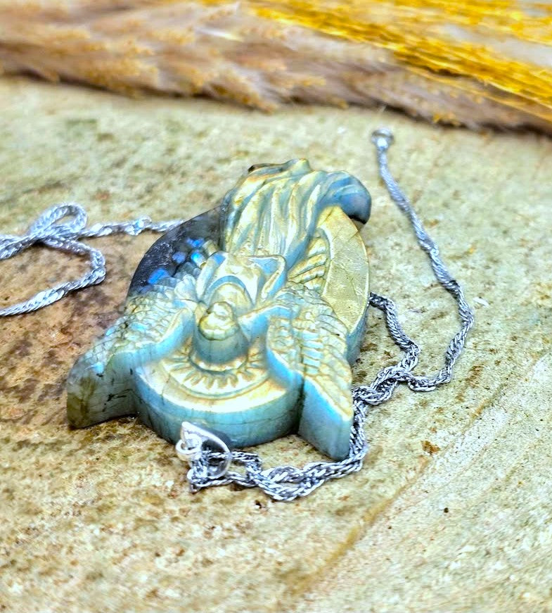 A carved pendant necklace with a blue-green stone featuring a winged figure design, resting on a textured stone surface with a silver chain.