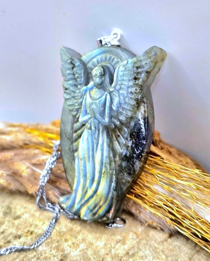 A silver-toned angel pendant necklace featuring a detailed angel figure with wings, set against a dark iridescent oval background, resting on a textured surface with dried grass.