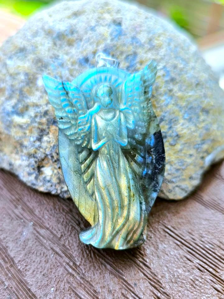 A carved angel pendant with a turquoise-blue and green iridescent finish, displayed on a wooden surface in front of a textured gray stone.