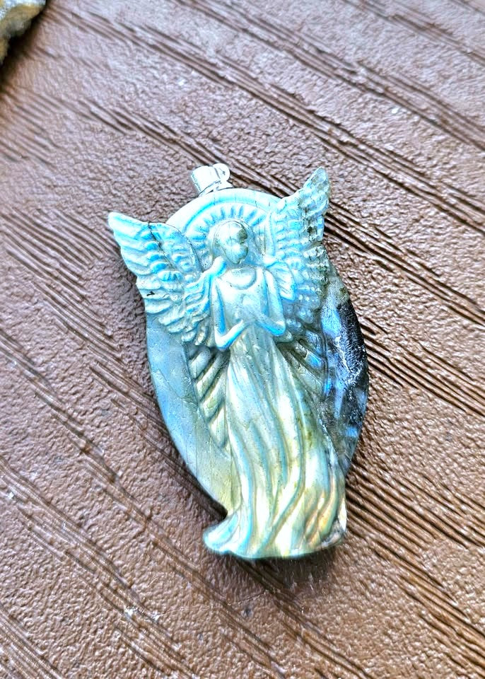 A carved angel pendant with iridescent blue, green, and gold tones, resting on a textured brown wood surface.