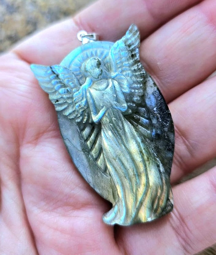 A hand holding a carved labradorite pendant featuring an angel figure with wings and a draped gown, showing iridescent blue, gold, and gray tones.