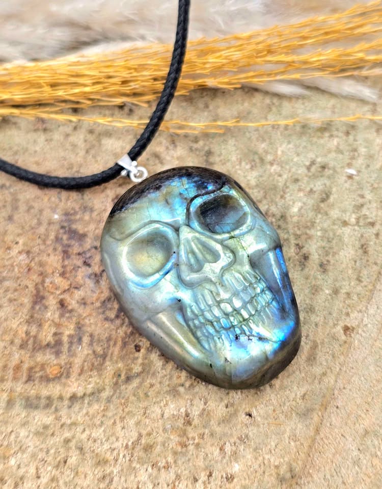 A skull-shaped pendant made of polished labradorite stone with a black cord necklace, displayed on a textured stone surface with dried grass.
