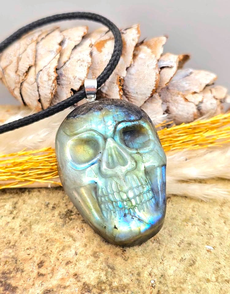 A skull-shaped pendant made of iridescent labradorite stone on a black cord necklace, displayed with a pine cone and dried grass on a textured surface.