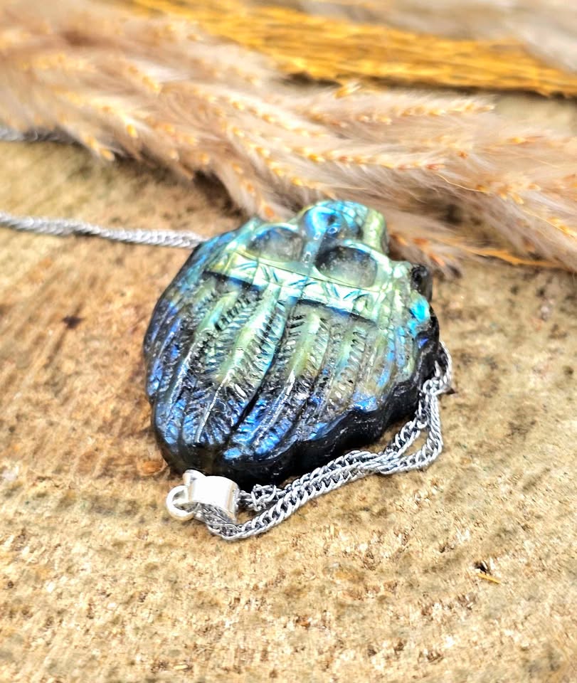 A carved skull-shaped pendant necklace with a vibrant blue and green iridescent finish, resting on a textured sandy surface with dried grass in the background.
