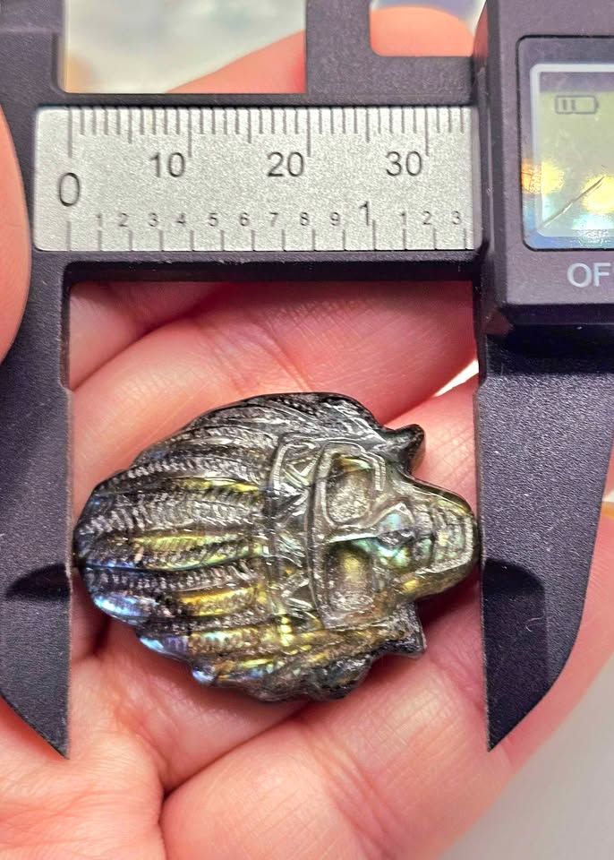 A close-up of a small iridescent metallic pendant or charm held in a person's hand under a digital caliper for scale. The object has a dark base with rainbow-like shifts including blue, green, and gold, and is photographed against skin and a black caliper.