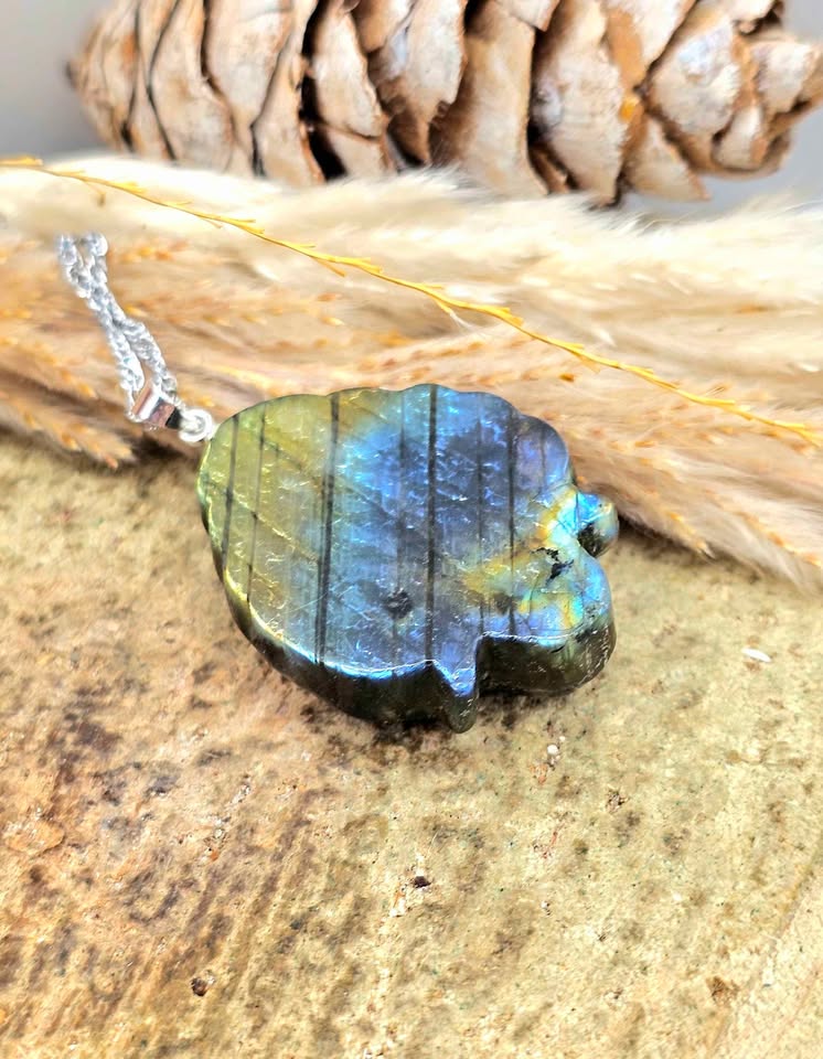 A polished labradorite stone pendant necklace with a silver bail, resting on a textured wooden surface with dried grass and a pine cone in the background.